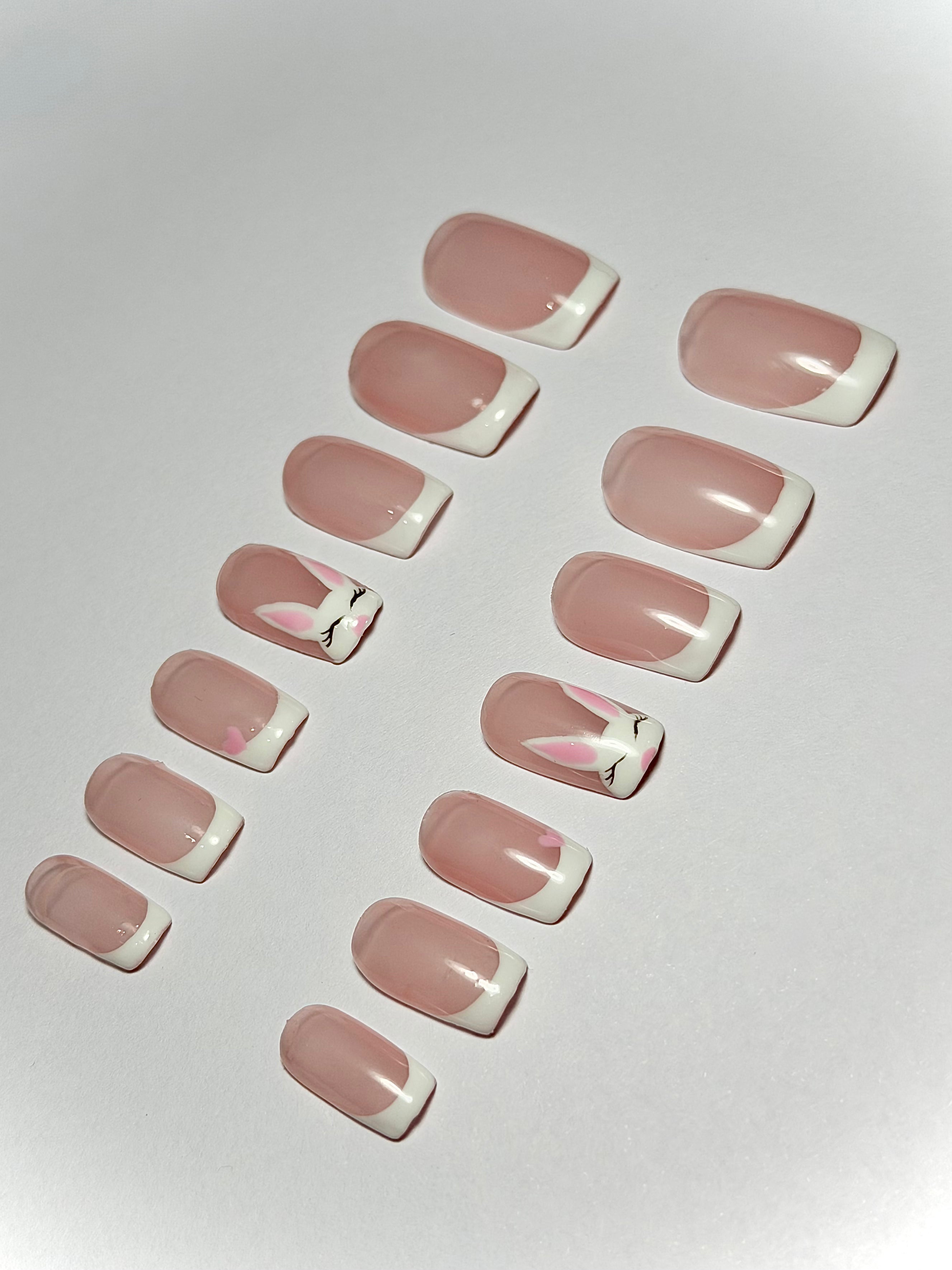 A set of press-on nails arranged on a light gray background. The nails have a glossy pale pink base with a white French tip and a small pink bunny face design on two of the nails.